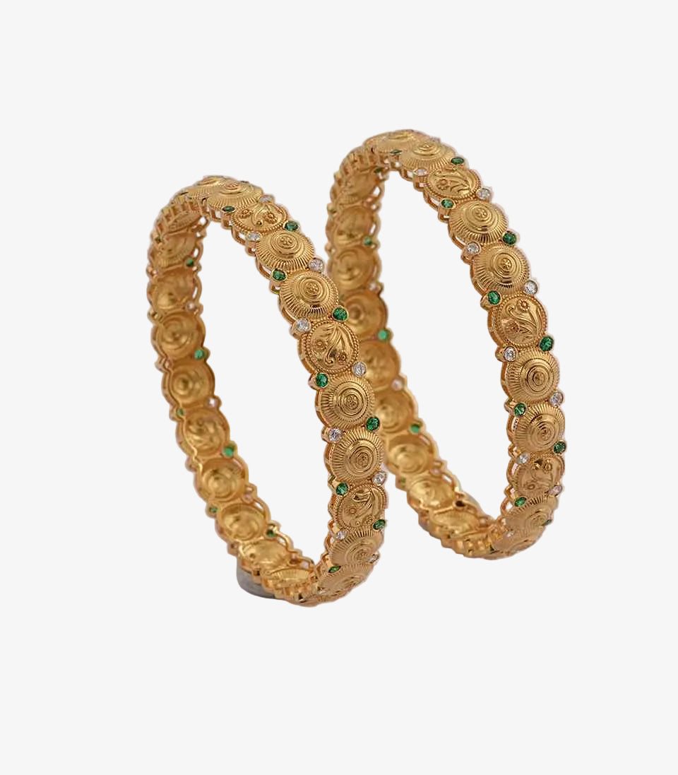 A pair of intricately designed gold bangles with circular engraved patterns, accented by small green and white stones, placed against a plain light background.